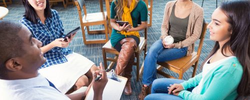 Diverse group of teen and young adult women are sitting in a circle during a meeting or support group therapy session. Young women are listening to mid adult African American male advisor or counselor. Women are holding digital tablets or coffee while listening.